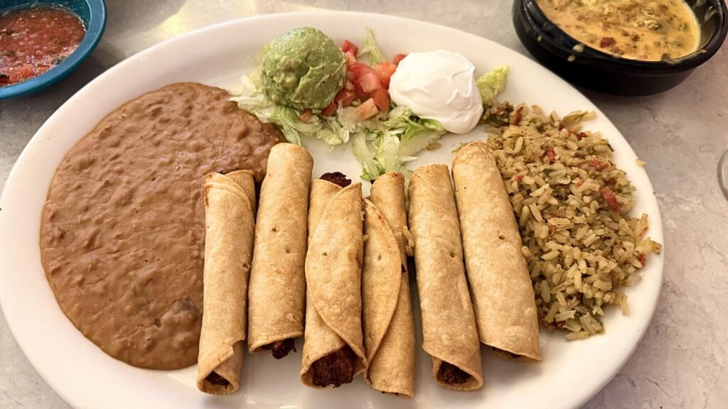 A plate of crispy taquitos served with refried beans, Mexican rice, guacamole, sour cream, and a lettuce and tomato salad.
