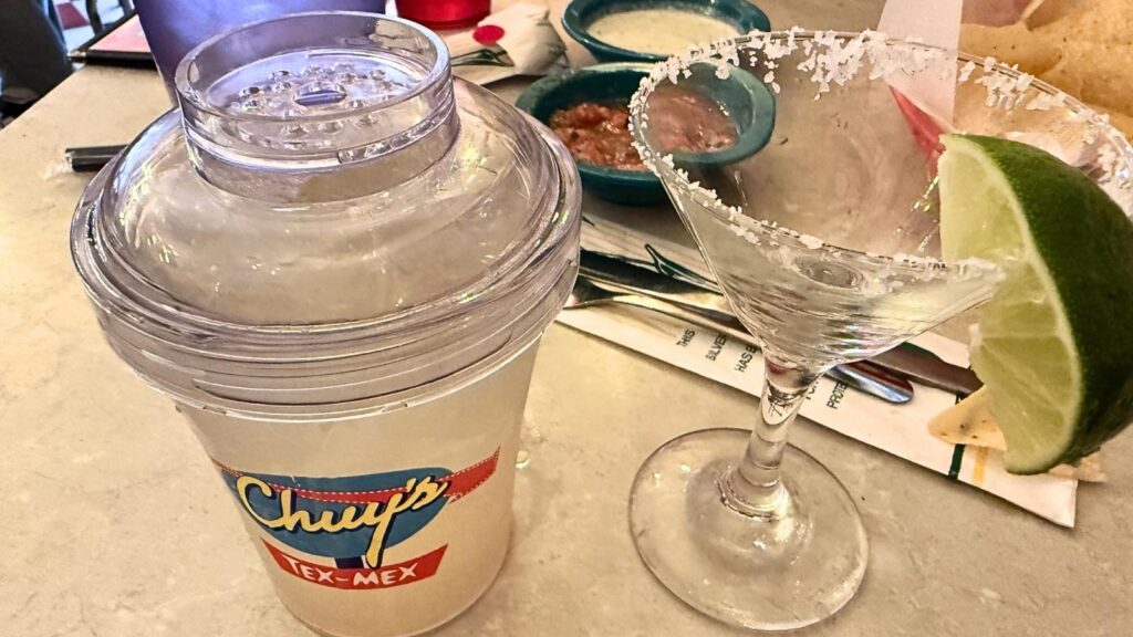 A frozen margarita in a Chuy's Tex-Mex shaker cup next to a salt-rimmed margarita glass with a lime wedge on a table.