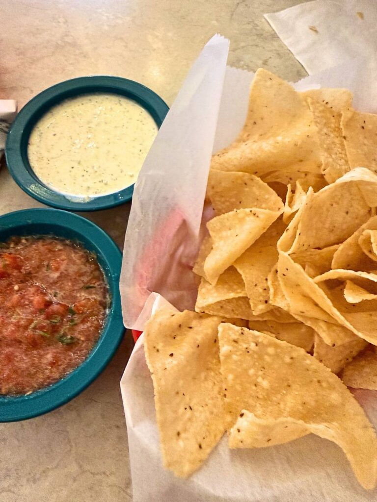 A basket of tortilla chips served with bowls of creamy jalapeño dip and fresh salsa on a restaurant table.